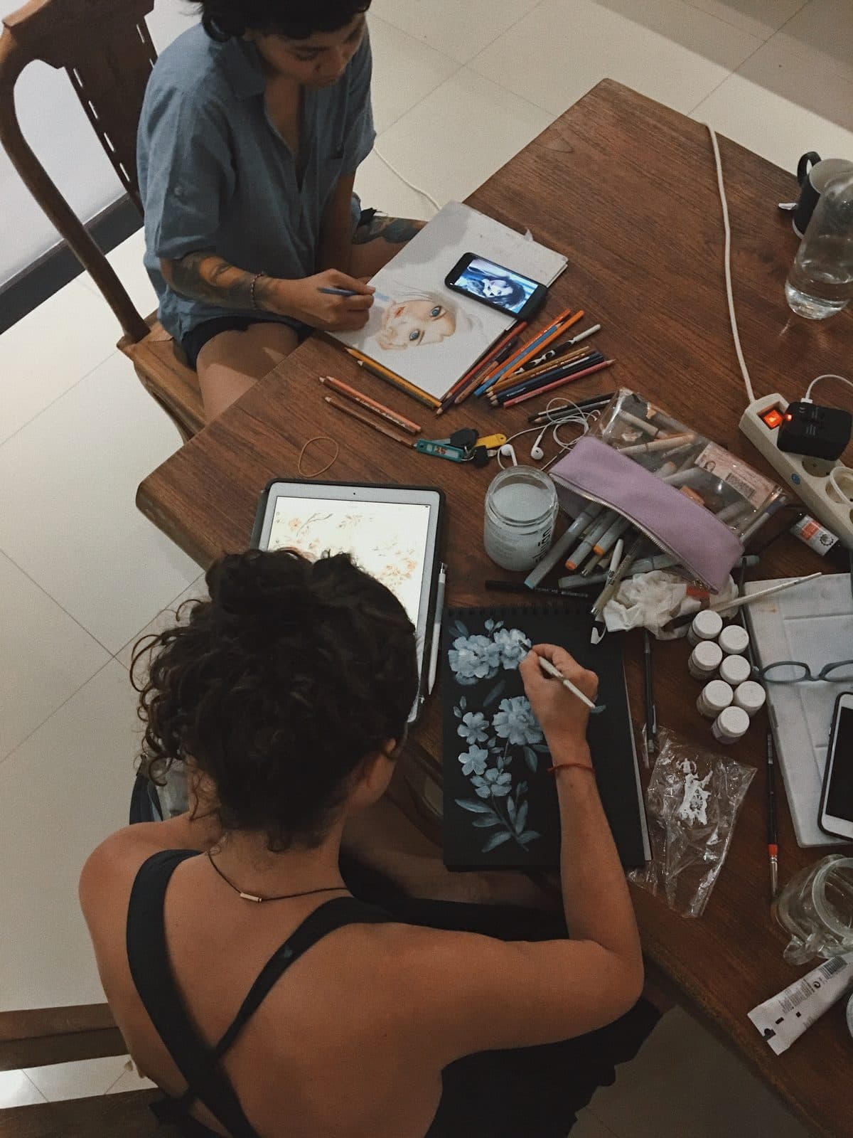 Two artists drawing and painting together at a table covered with pencils and supplies
